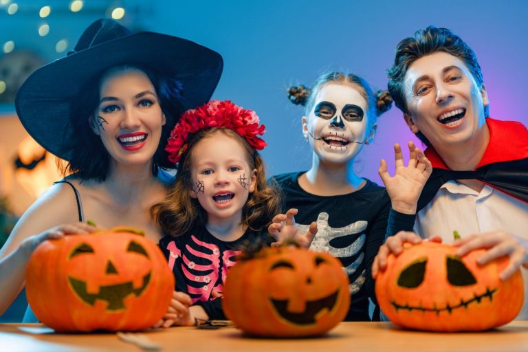 Family dressed in Halloween costumes, safely trick-or-treating at a well-lit home decorated with pumpkins and spooky decor